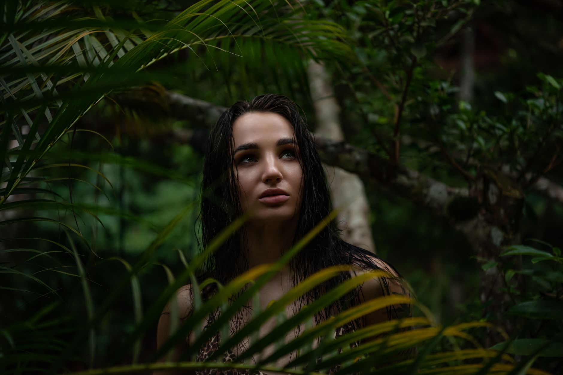 A woman stands amidst tropical foliage, looking thoughtfully upward in a jungle environment.