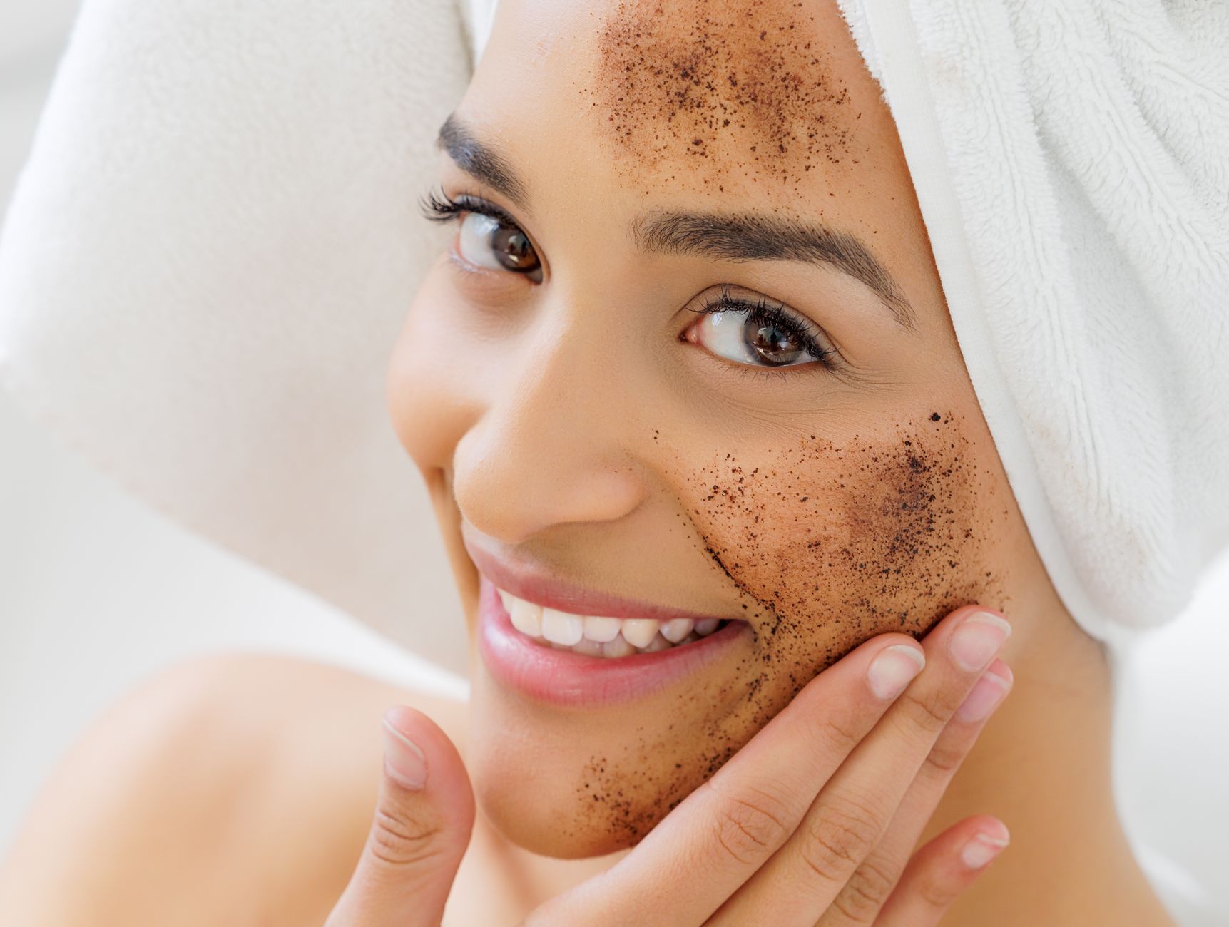 Smiling woman with towel on her head applying a natural exfoliating face mask. Skincare and self-care concept.