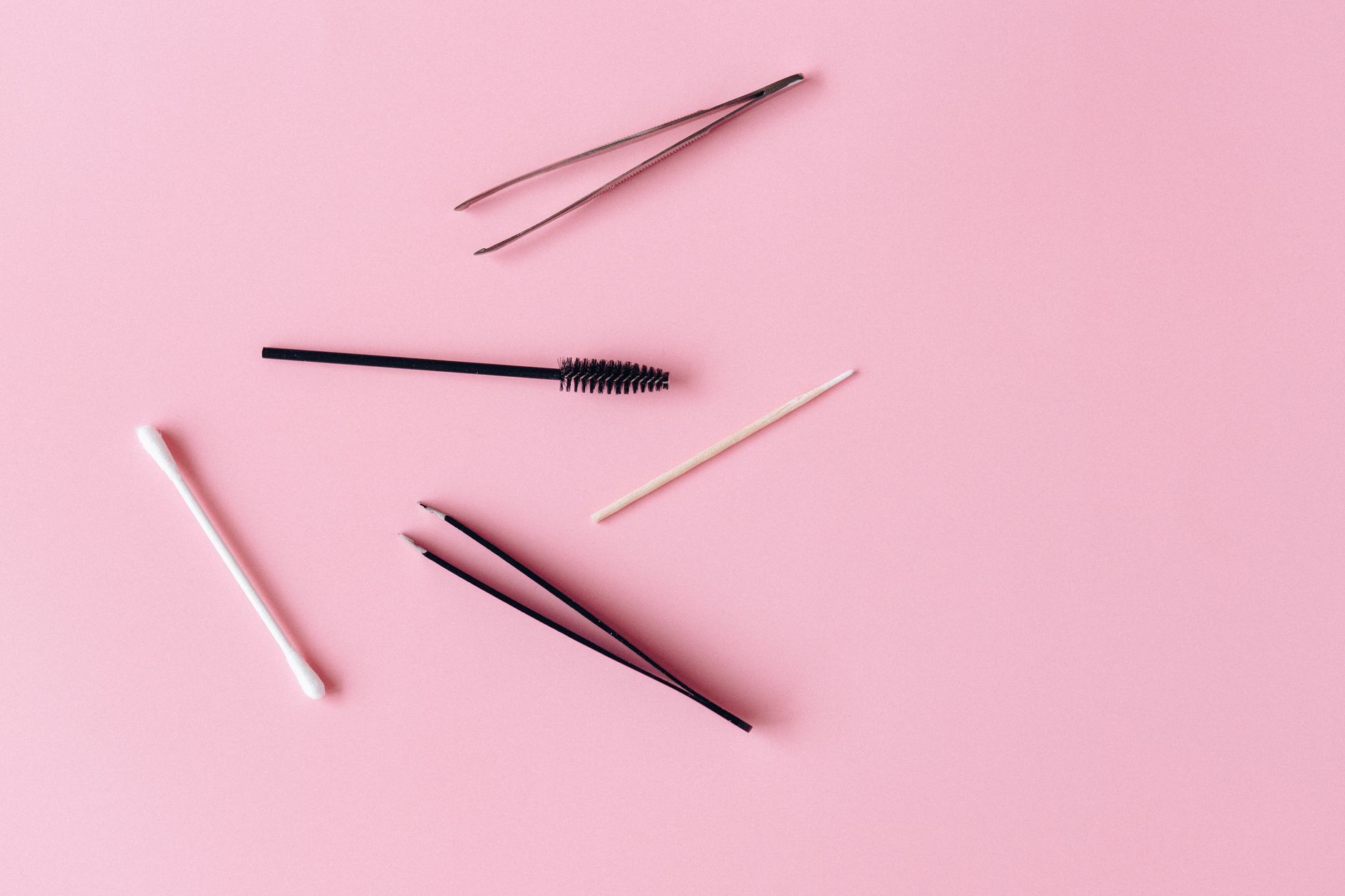 Colorful flat lay of makeup tools featuring tweezers and brushes on a pink background.