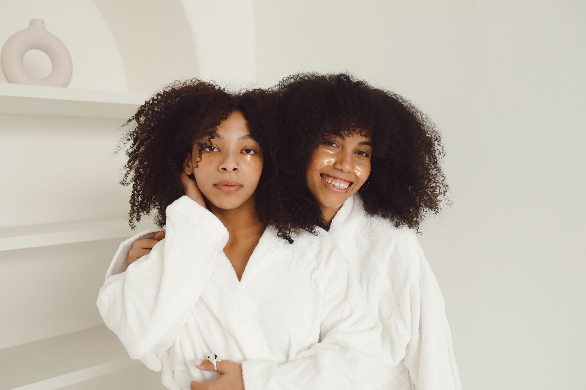 Two women enjoying a skincare routine with facial patches and bathrobes, embracing each other joyfully indoors.