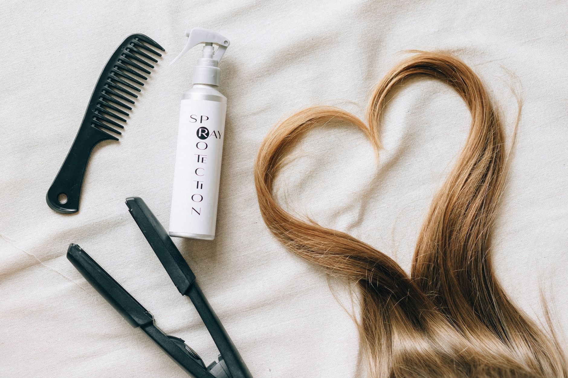 Flat lay of hair styling tools on a bedsheet with heart-shaped hair strand.