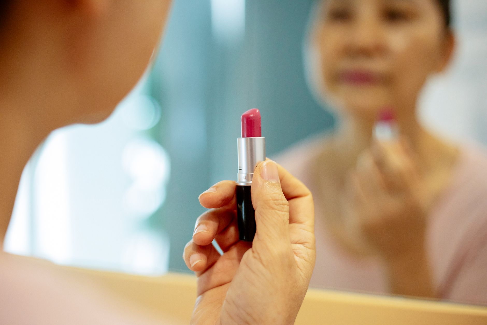Confident middle age female in casual outfit with purple lipstick looking at mirror reflection in bright apartment