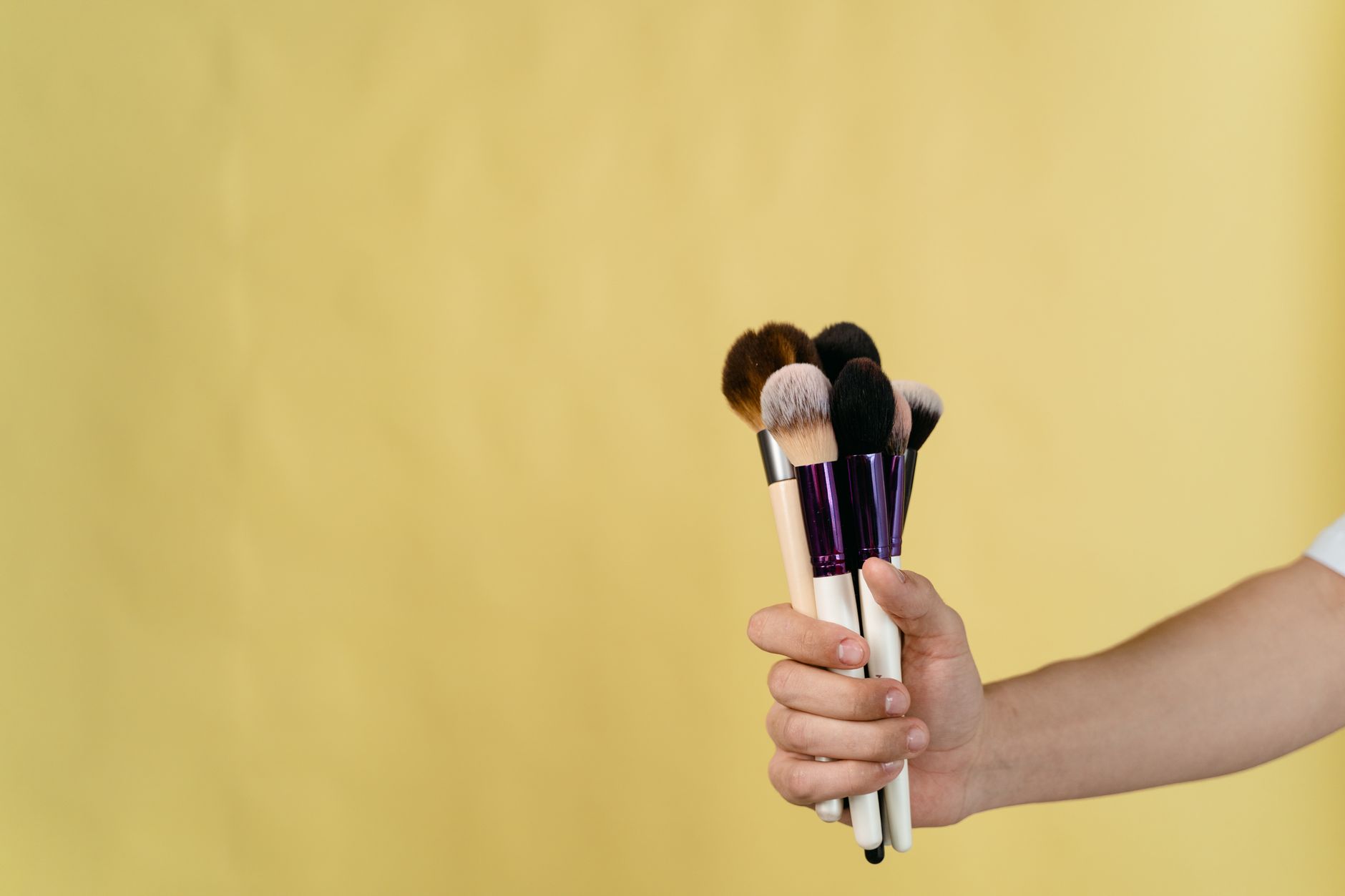 Close-up shot of a hand holding a set of makeup brushes against a pale yellow background.