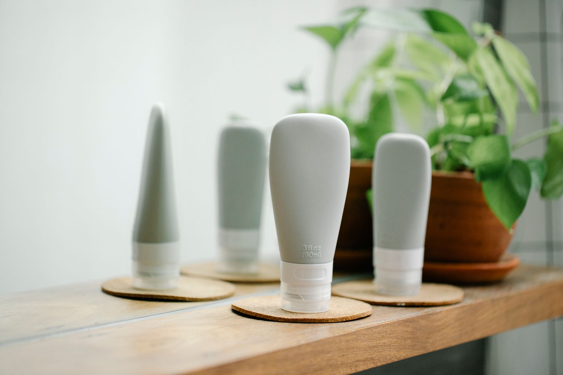 Gray tubes with skincare products placed on coasters on wooden shelf near mirror in light room with green potted plant