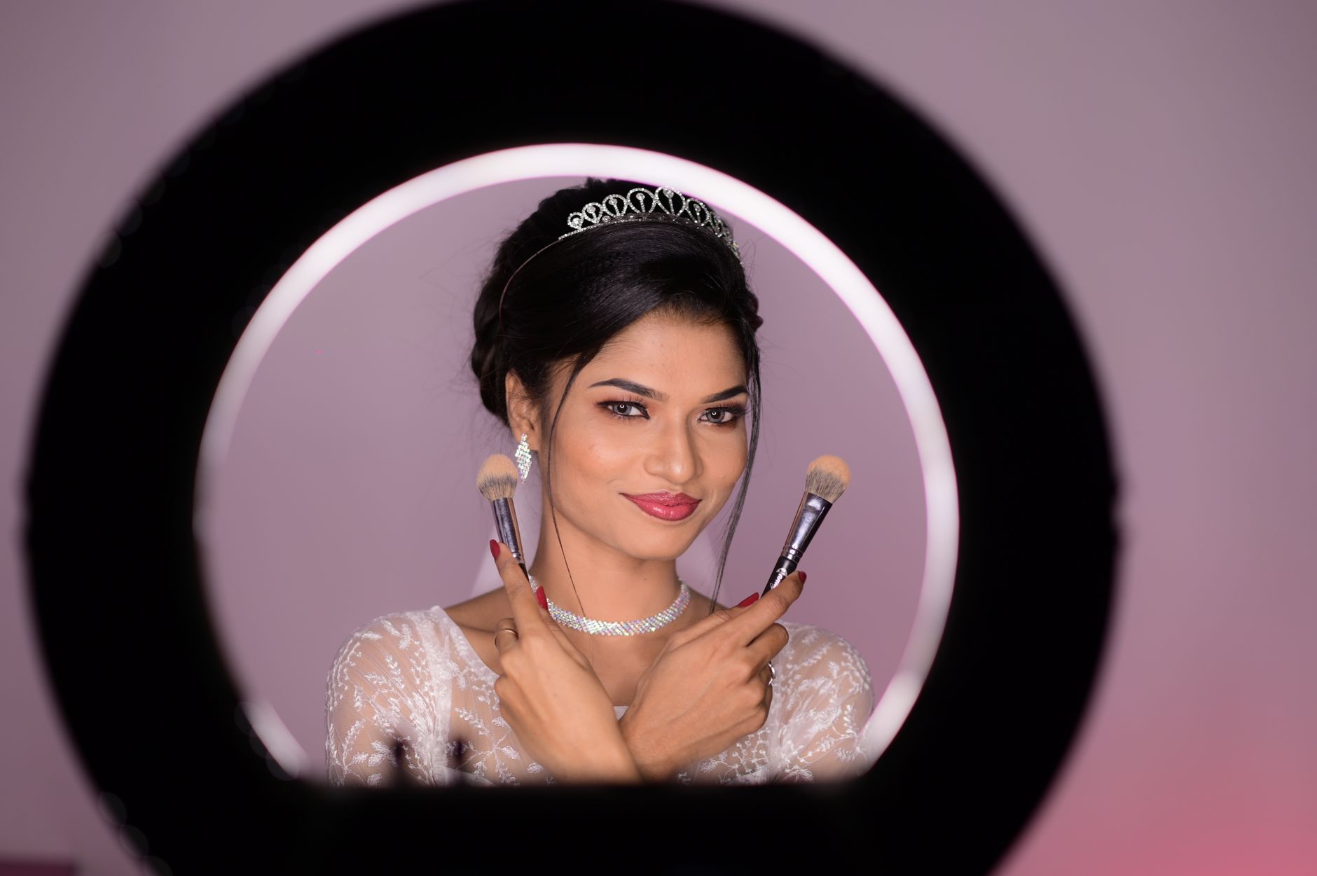 A glamorous woman wearing a tiara poses with makeup brushes in a studio setting under professional lighting.
