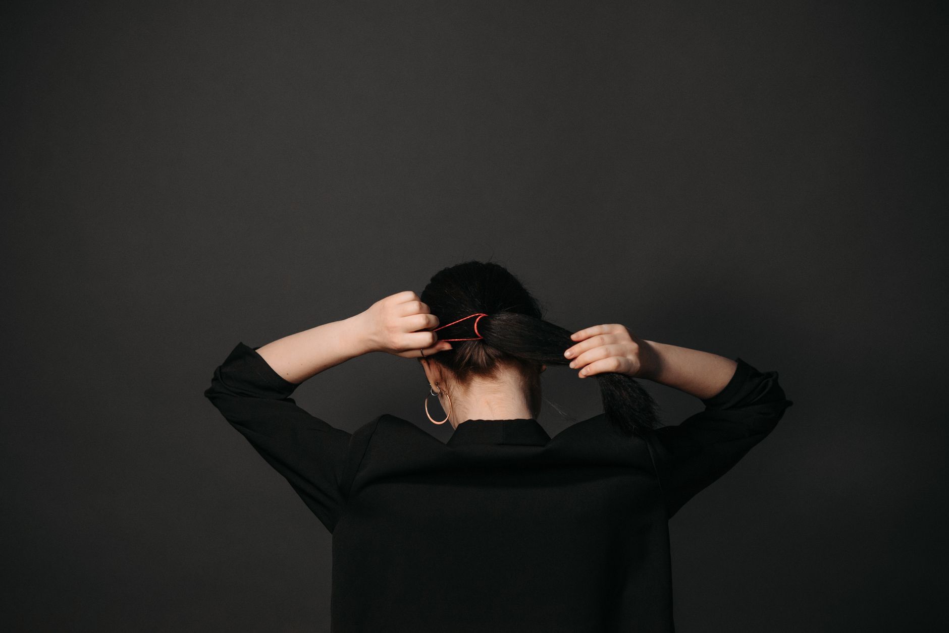 Woman tying her hair into a ponytail against a dark background, focusing on hairstyle and casual elegance.