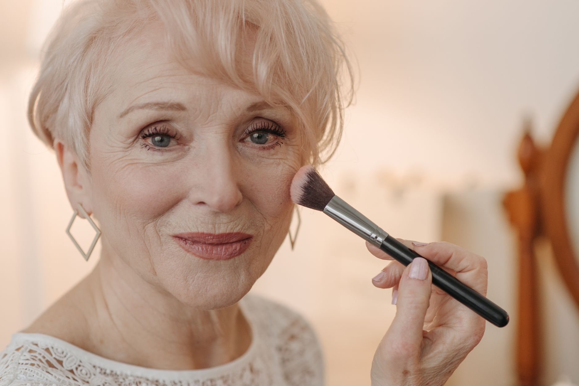 A senior woman with short hair applies blush with a smile indoors.