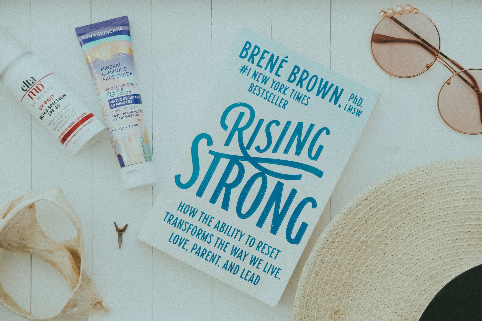Flat lay of summer essentials including "Rising Strong" book, sunscreen, sunglasses, and straw hat on white planks.