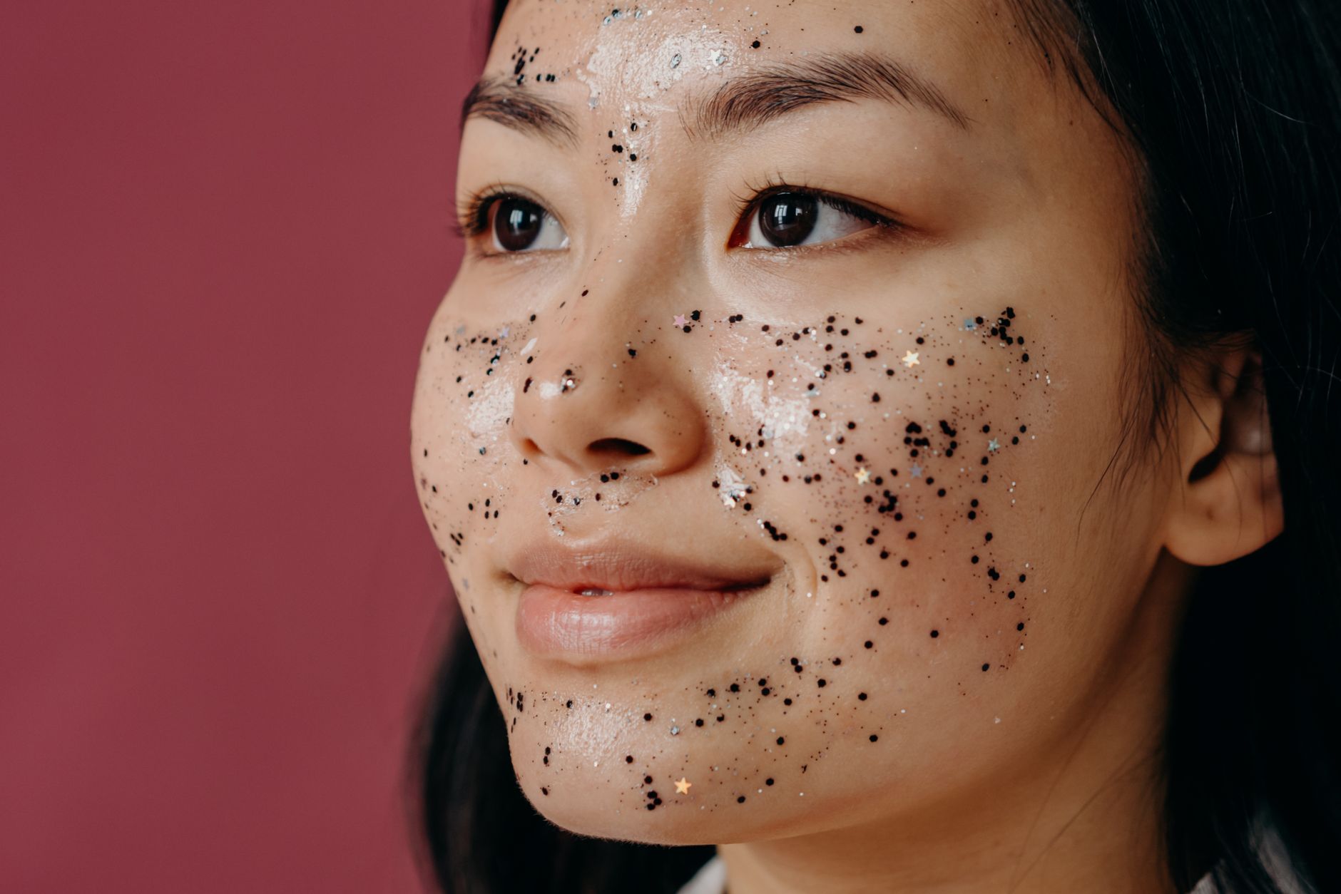 Asian woman with glitter on face smiling against a pink background. Studio portrait.
